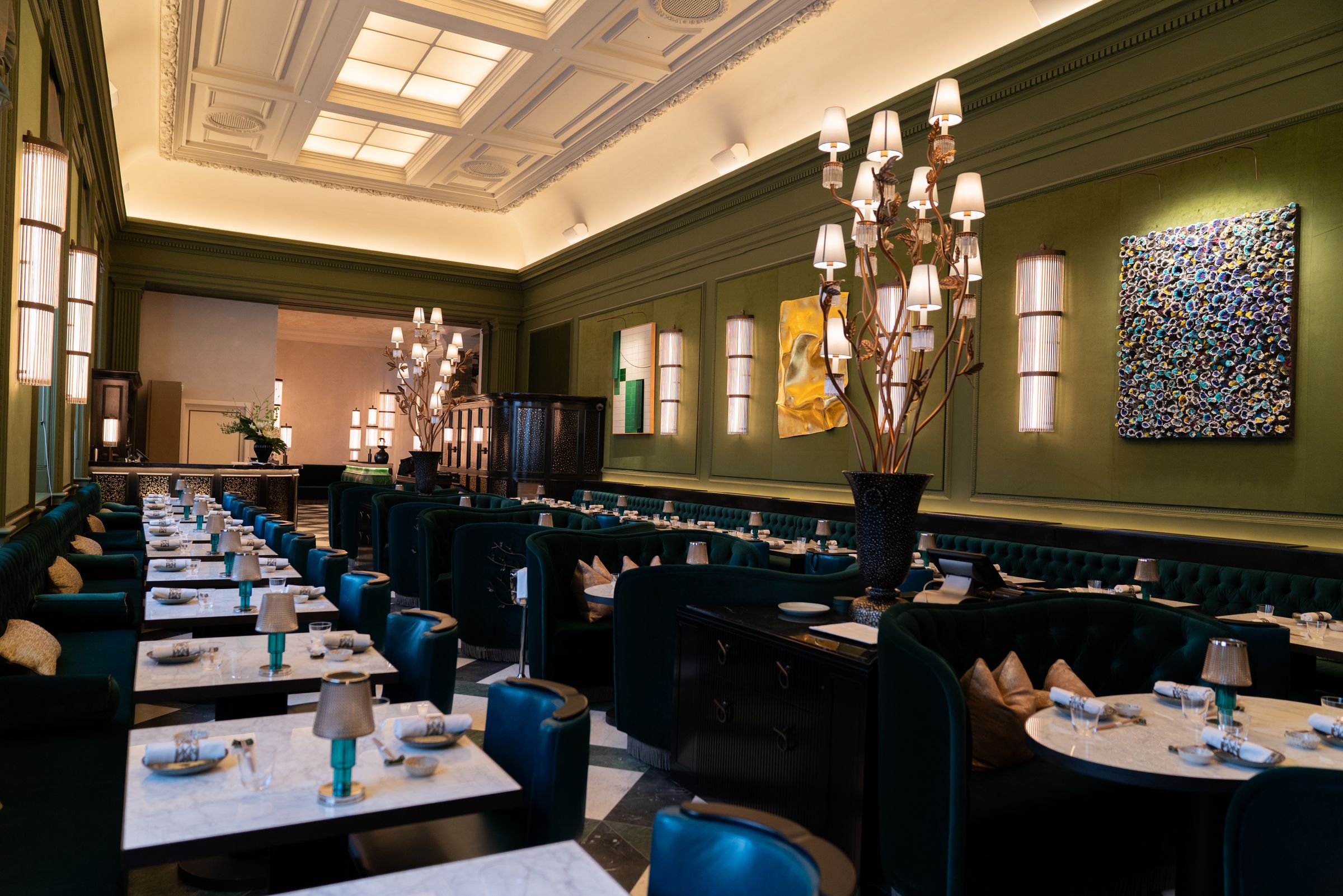 A long green-panelled dining room under a coffered ceiling, with a tall branched candelabrum, marble tables and blue chairs. Three works hang on the right-hand wall: a green and white tiled panel, a sheet of gold and a panel of dense rosettes.