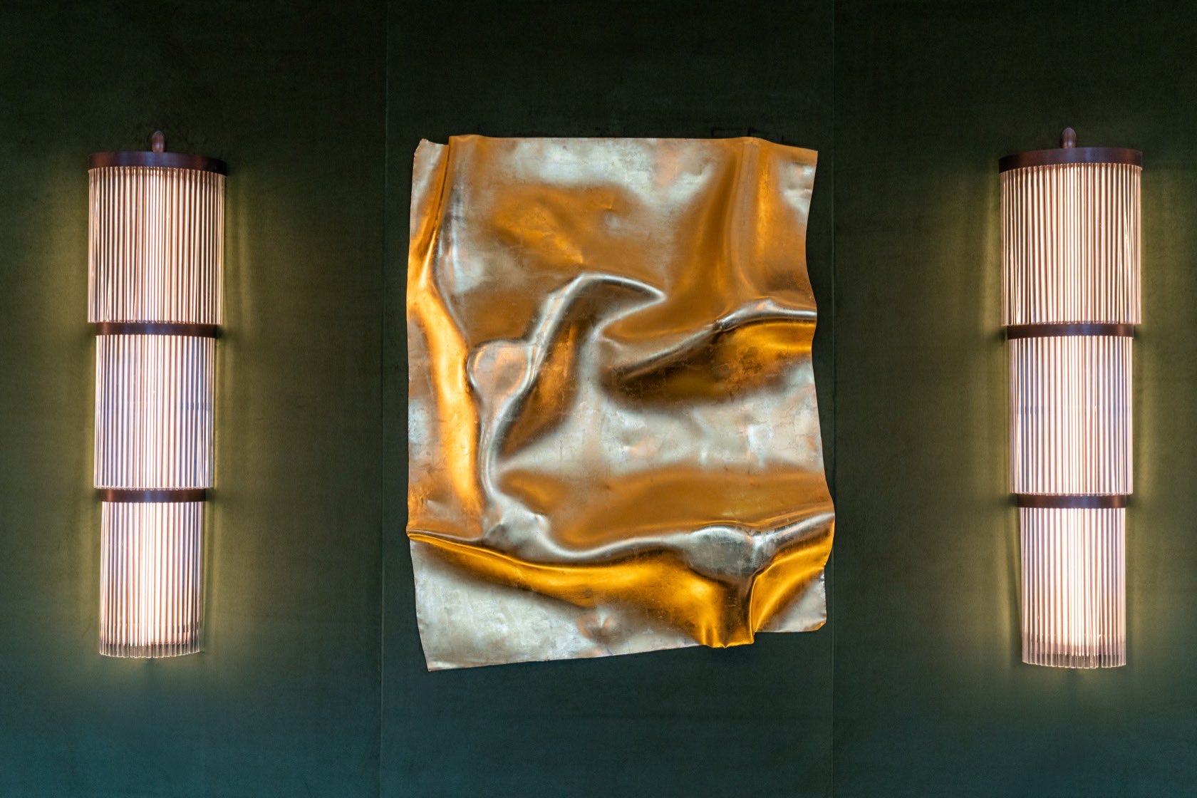 A folded gold sheet mounted on a dark green wall, its creases catching gold and silver light, flanked by two tall ribbed wall lights.