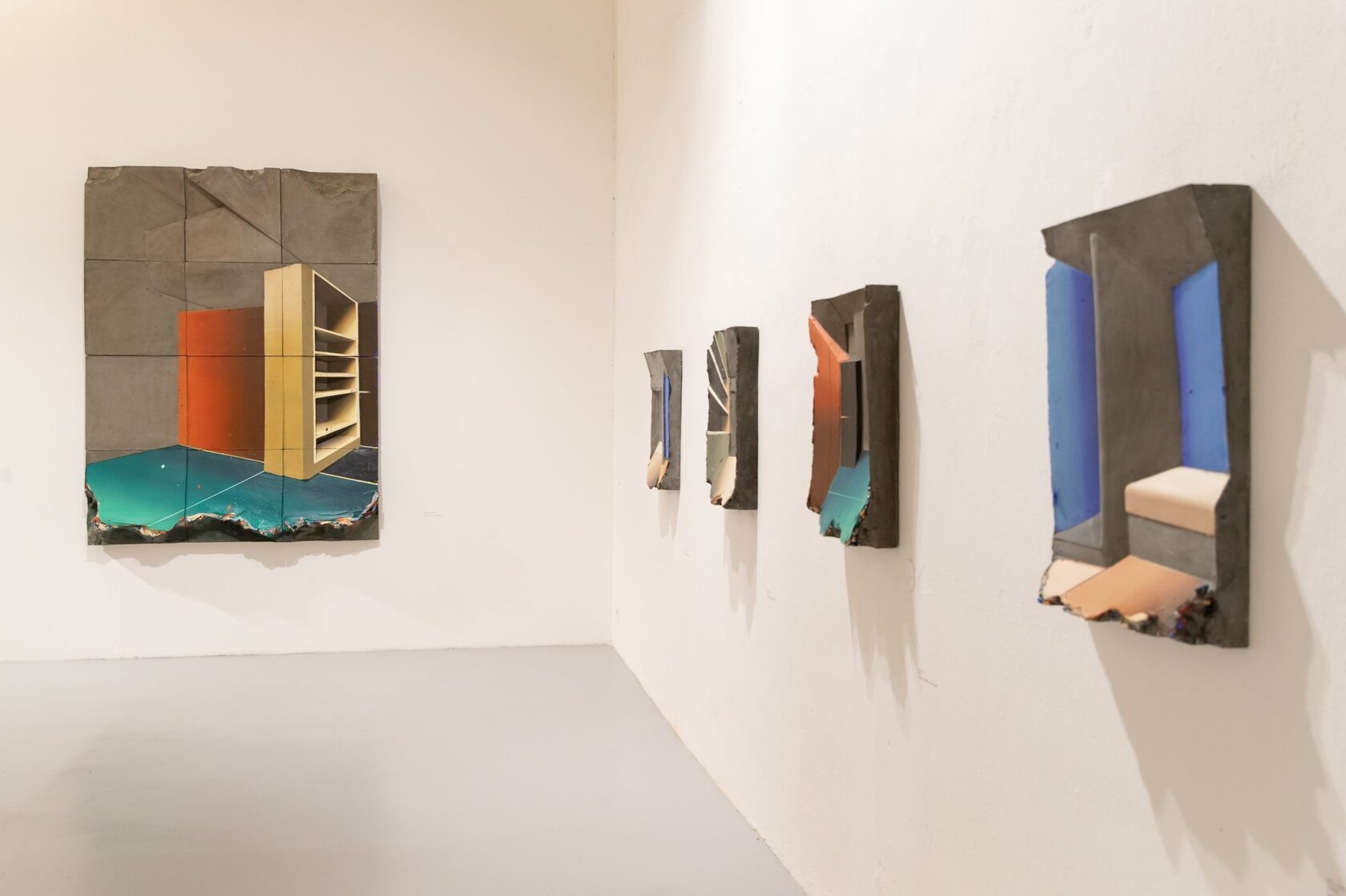Five painted relief panels across a corner of a white gallery: a large one on the back wall and four spaced along the wall at the right.